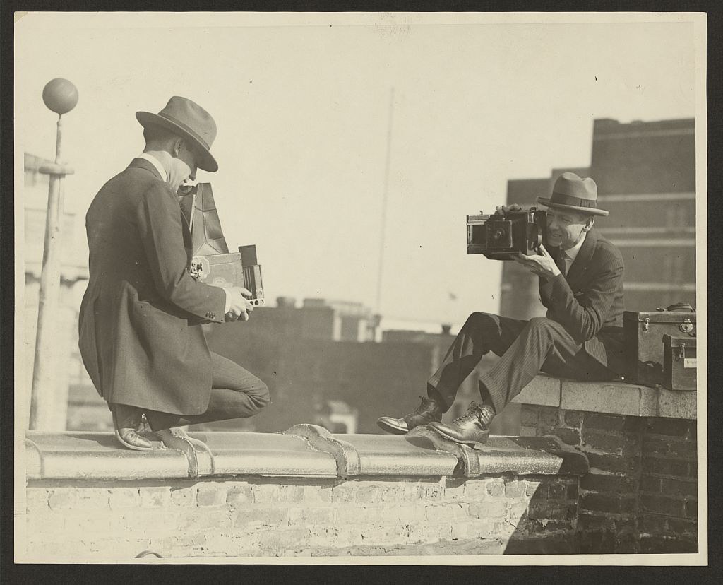 [Two photographers taking each others' picture with hand-held cameras while perched on a roof] (LOC)